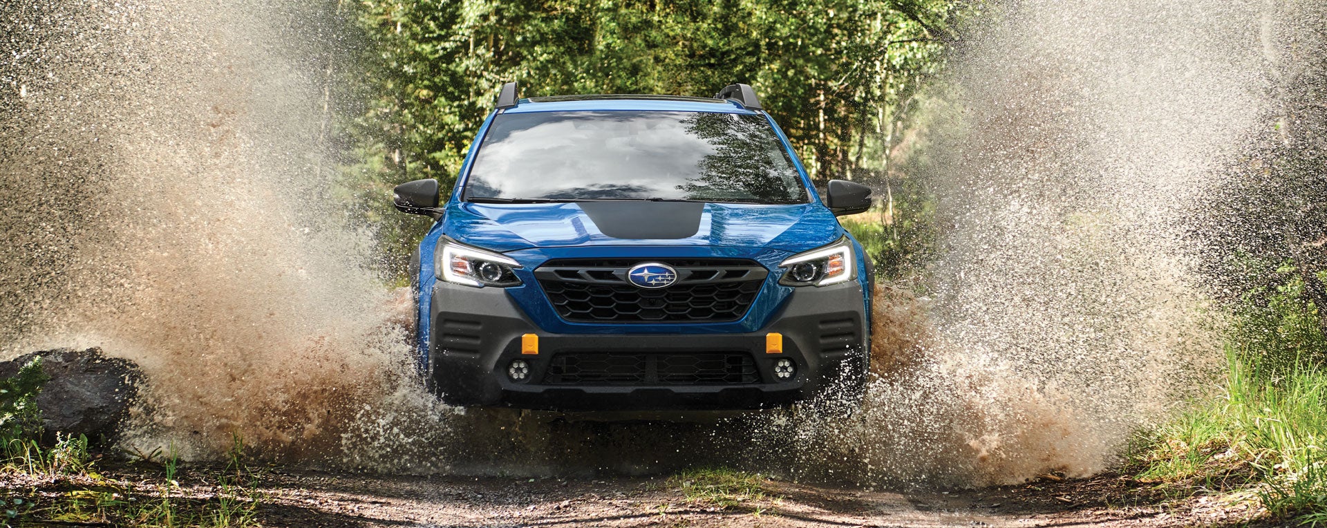 Front view of a blue Subaru outback_wilderness wading through water that splashes high on both sides toward green trees.