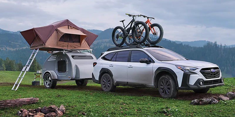 White Subaru Outback towing a small camper with a rooftop tent and bicycles on the roof in a grassy campsite.