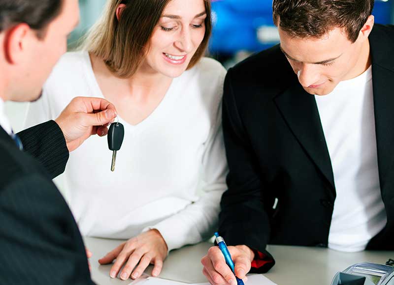 Couple signing car purchase paperwork while dealer hands over car key.